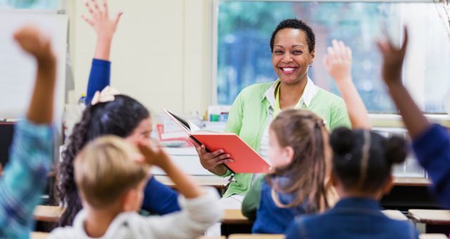 Smiling teacher reading to class from a book while students listen, some raising their hands.