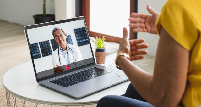 Person sitting at table looking at a laptop screen while gesturing with their hands, talking to a doctor conducting a telehealth appointment.