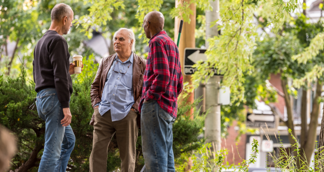Three neighbors standing on a sidewalk by a fence talking.