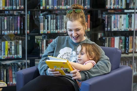 A mother sits in a comfortable chair at the Library reading books aloud to the child sitting in her lap.