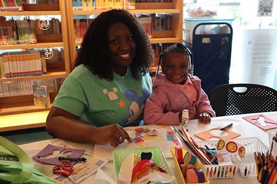 A small child and her caregiver sitting at a craft table working with paper shapes and glue.