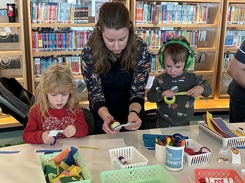 An adult helps two children make a craft using scissors and colorful tape.