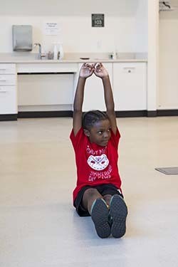 A young child sitting on the floor with their legs straight in front of them and their arms stretched overhead.